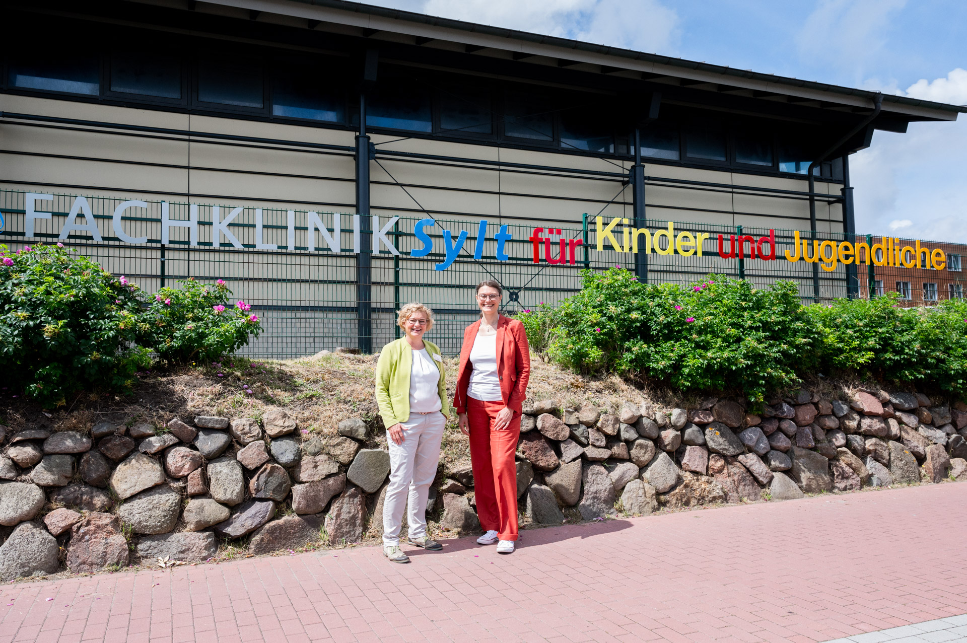 Foto der Klinikleitung Dr. Ines Gellhaus und Saskia Louwers vor dem Namensschild der Fachklinik Sylt für Kinder und Jugendliche